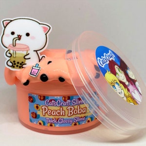 Pu&ograve; includere: Un contenitore di plastica trasparente riempito di slime color pesca con palline di boba nere. Il contenitore ha un'etichetta che recita "CatsCraft Slime Peach Boba Thick Glossy Slime". Un cartone animato di un gatto bianco con una tazza di boba tea rosa &egrave; sul coperchio.
