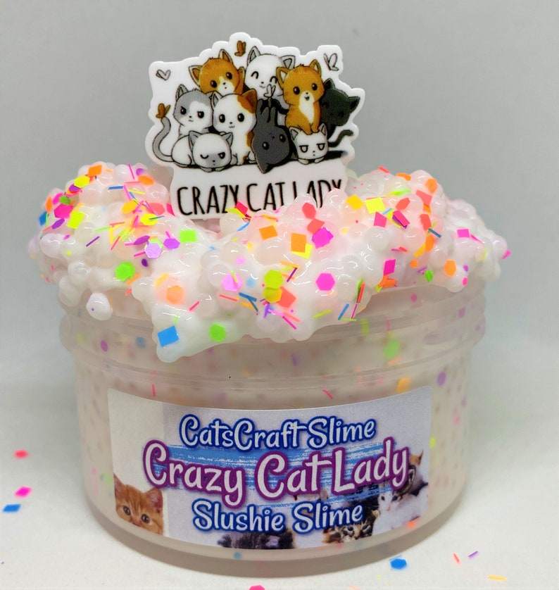 May include: A clear jar filled with white slushie slime with colorful glitter and sprinkles. The jar has a label that reads "CatsCraft Slime Crazy Cat Lady Slushie Slime". A charm with a group of cartoon cats is on top of the jar.