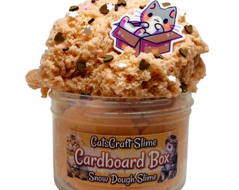 May include: A clear plastic container filled with a light orange snow dough slime. The slime is topped with brown and white sprinkles and a small cartoon cat figurine sitting in a cardboard box.