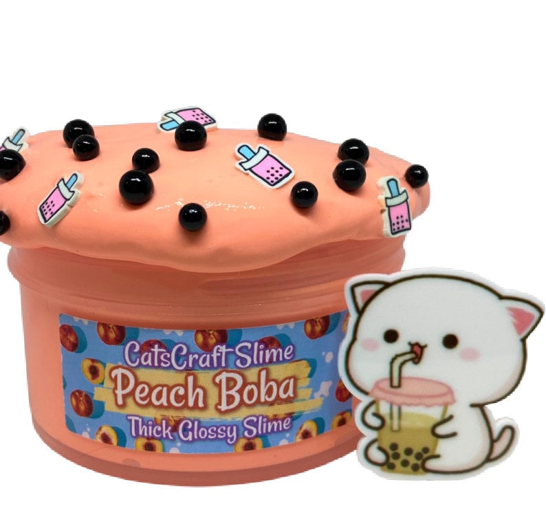 Pu&ograve; includere: Slime Peach Boba in un contenitore trasparente con un designo di pesca e boba. Lo slime &egrave; ricoperto da palline di boba nere e ciondoli a tema pesca. Il contenitore ha un'etichetta che recita "CatsCraft Slime Peach Boba Thick Glossy Slime". Un ciondolo di gatto bianco con una bevanda boba si trova sul lato destro dell'immagine.