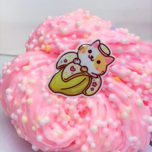 May include: Pink slime with white and yellow sprinkles and a small cartoon sticker of a cat wearing a halo and holding a banana.