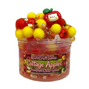 May include: A clear plastic jar filled with red and yellow slime, topped with decorative elements. The slime is adorned with red and yellow pom-poms, a red apple-shaped charm, and small leaf accents. The jar has a label that reads "CatsCraft Slime Cottage Apples PomPom Clear Slime."