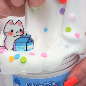 May include: White slime with colorful sprinkles and a cartoon cat holding a carton of milk. The slime is in a clear jar with a label that reads "CatsCraft Slime Meow Milk Thick Glossy Slime".