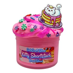 May include: A pink slime in a clear plastic container with a label reading CatsCraft Slime Kitty Shortstack Bread Slime. The top is decorated with pink slime swirls, colorful circular candy-like charms, and a cat on pancakes topper.