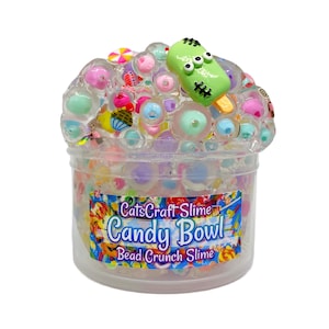 May include: A clear plastic container filled with clear slime containing various colorful beads and small candy-themed charms, including a green three-eyed popsicle charm. The label reads CatsCraft Slime Candy Bowl Bead Crunch Slime.