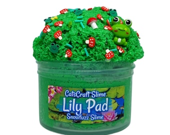 May include: A clear plastic jar filled with green slime, decorated with green sprinkles, red and white mushroom-shaped ornaments, and a small green frog figure. The jar is labelled "CatsCraft Slime Lily Pad Snowfuzz Slime."