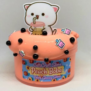 Pu&ograve; includere: Slime alla boba di pesca con palline di boba nere e ciondoli di boba rosa. Lo slime &egrave; in un barattolo trasparente con un'etichetta pesca e bianca che dice "CatsCraft Slime Peach Boba Thick Glossy Slime". Un gatto bianco con orecchie rosa e una bevanda boba rosa &egrave; sopra lo slime.