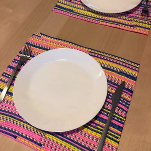 May include: Two white plates on colorful woven placemats. The placemats have a striped pattern in pink, yellow, and blue. A fork and knife are placed on the mats. The setting is on a light wood table.