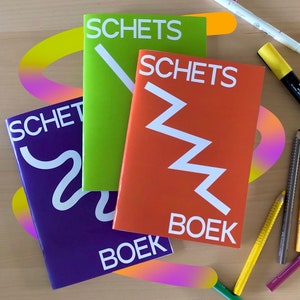 May include: Three sketchbooks in purple, green, and orange, each with the word "SCHETS" and "BOEK" in white. The orange book features a white zigzag design. Colored pencils and markers are scattered around the books on a wooden surface.