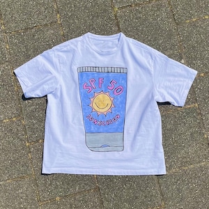 May include: White t-shirt featuring a cartoon illustration of a sunscreen tube with a smiling sun and the text "SPF 50 SUNSCREEN" in blue and pink. The shirt is a casual, short-sleeved design.
