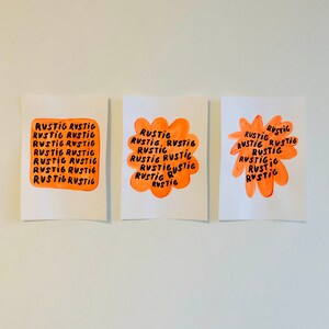 May include: Three white paper prints with orange and black text. The word "RUSTIG" is repeated in a rectangular, flower, and starburst shape. The prints are hung on a white wall.