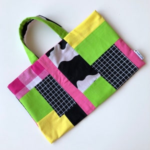 May include: A patchwork tote bag with a lime green handle and a rectangular body. The body is composed of various fabric patterns, including pink plaid, black and white grid, cow print, and solid blocks of pink, yellow, and green.