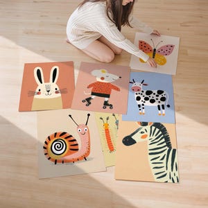 May include: A collection of colorful animal illustrations on square cards. The images include a rabbit, mouse on roller skates, butterfly, cow, snail, caterpillar, and zebra. The cards are arranged on a light-colored wooden floor.