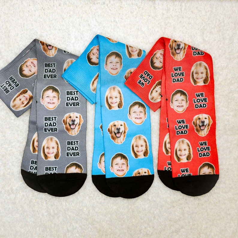 May include: Three pairs of socks with different designs. The socks are all white with a black cuff. The first pair has a blue background with a pattern of dogs, children, and the text "Best Dad Ever". The second pair has a red background with a pattern of dogs, children, and the text "We Love Dad". The third pair has a gray background with a pattern of dogs, children, and the text "Best Dad Ever".