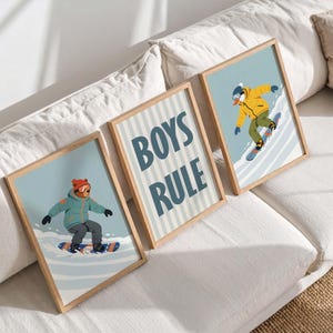 May include: Three framed art prints featuring illustrations of snowboarders and the text "BOYS RULE." The prints have light blue backgrounds and wooden frames. The snowboarders are wearing winter gear in shades of blue, yellow, and orange.