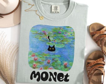 May include: A light blue t-shirt with a black cat swimming in a painting of water lilies. The painting is a recreation of Claude Monet's famous work. The text "MONET" is printed in black below the painting.