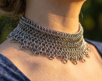 May include: A silver chainmail choker necklace. The necklace is made of small, interlocking metal rings and sits around the neck. The model is wearing a dark blue velvet top.