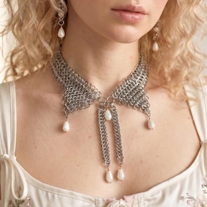 May include: A silver chainmail collar necklace with dangling white pearl accents. The necklace is paired with matching pearl drop earrings. The model is wearing a white floral embroidered top.