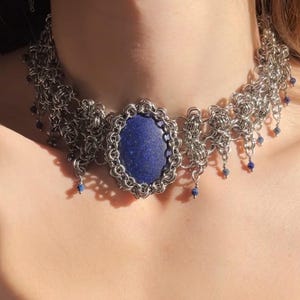 Peut inclure: Un collier ras-du-cou en cotte de mailles argentée avec une pierre ovale en lapis-lazuli bleu profond sertie dans un cadre en métal décoratif. Le collier est orné de petites perles bleues suspendues le long du bord inférieur des maillons.