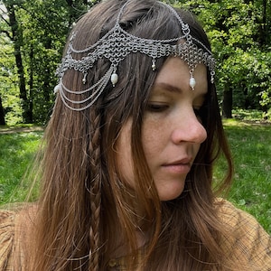 May include: A silver chainmail headpiece with pearl accents. The headpiece is worn by a person with long brown hair styled in braids.
