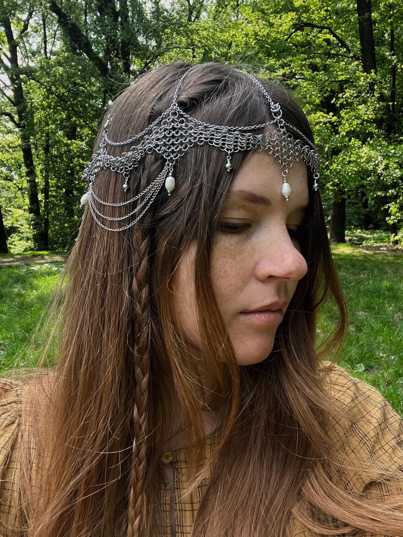 May include: A silver chainmail headpiece with pearl accents. The headpiece is worn by a person with long brown hair styled in braids.