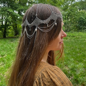 May include: A silver chainmail headpiece with pearl accents. The headpiece is worn by a person with long brown hair. The person is wearing a brown and black plaid shirt.