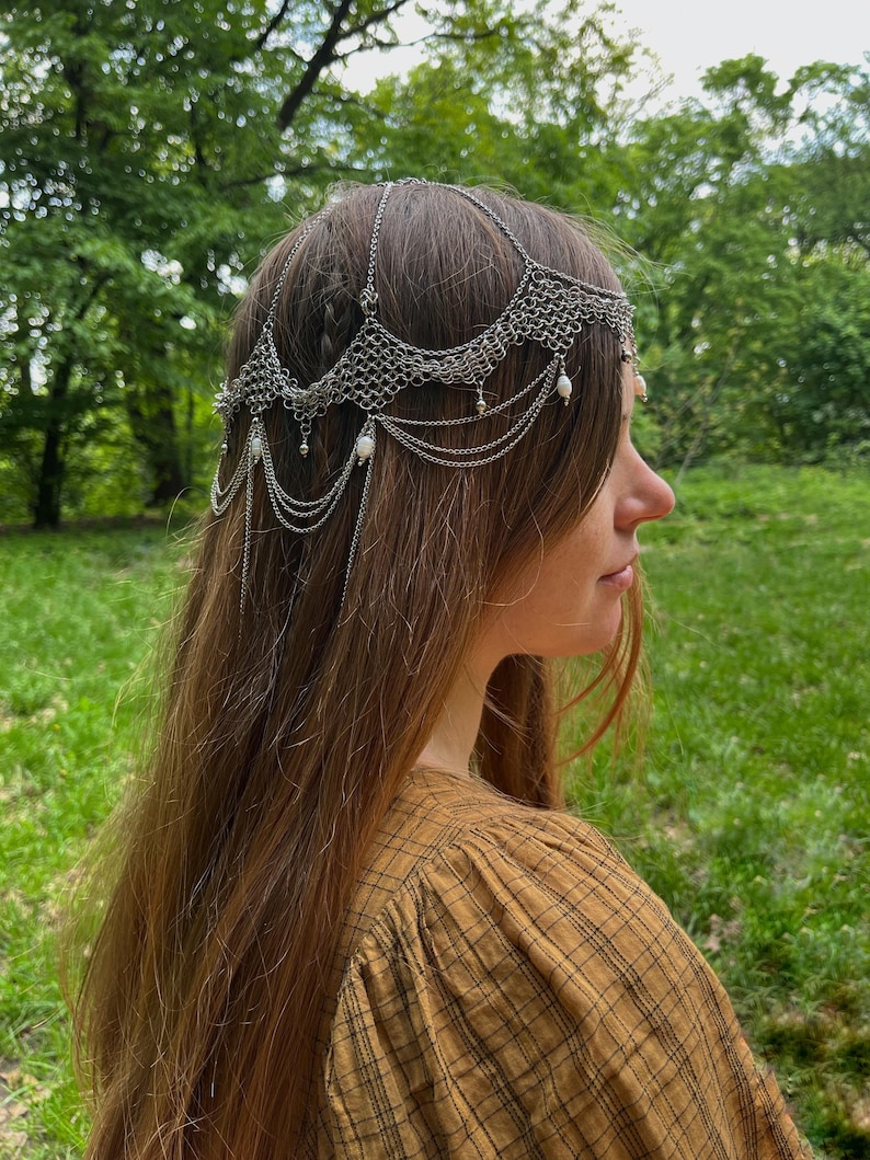 May include: A silver chainmail headpiece with pearl accents. The headpiece is worn by a person with long brown hair. The person is wearing a brown and black plaid shirt.