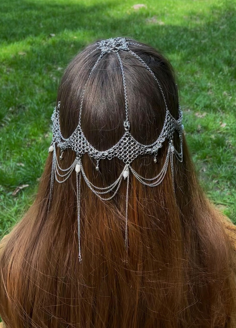 May include: A silver chainmail headpiece with dangling chains and white beads. The headpiece is worn on a person with long brown hair.