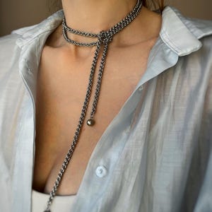 May include: A silver-toned chain necklace with a knotted design and two dangling strands. Each strand ends with a small, round bead. The necklace is worn over a light blue button-down shirt.