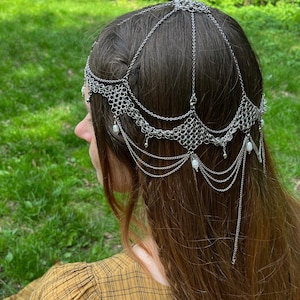 May include: A silver chainmail headpiece with pearl accents. The headpiece is worn by a person with long brown hair. The headpiece is made of metal chain links and has a delicate, intricate design.