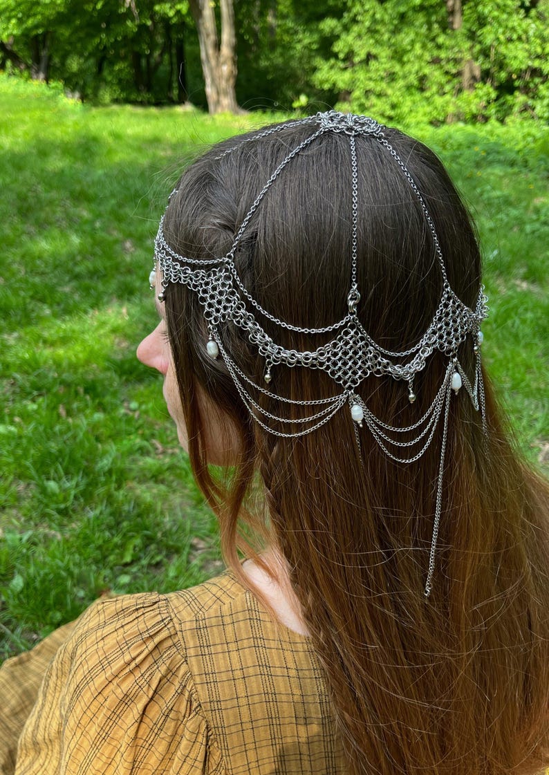May include: A silver chainmail headpiece with pearl accents. The headpiece is worn by a person with long brown hair. The headpiece is made of metal chain links and has a delicate, intricate design.