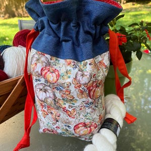 May include: A white drawstring bag with a fall-themed print of pumpkins, leaves, and berries. The bag has a blue denim top and red drawstrings. A ball of white yarn is next to the bag.