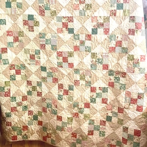May include: A handmade quilt with a patchwork design. The quilt features squares of fabric in various patterns and colors, including red, green, and cream. The quilt is made of soft fabric and has a quilted pattern throughout.
