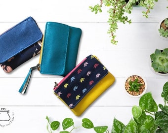 May include: Three small fabric wallets with different patterns and colors. The wallets are folded closed and have a zipper closure. The wallets are on a white wooden surface with green plants in pots.