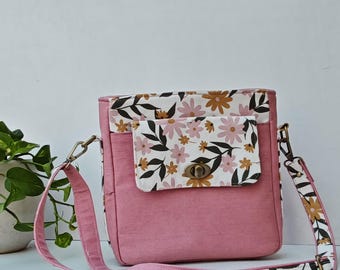 May include: A pink crossbody bag with a floral print flap and strap. The bag features a rectangular shape with a metal clasp closure. The strap and flap have a white background with pink, brown, and black floral patterns.