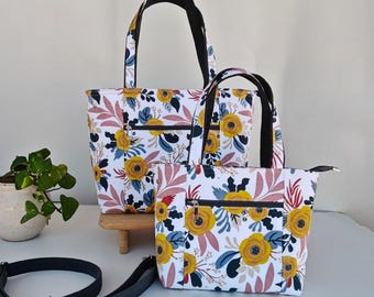 May include: Two tote bags featuring a white background with a floral pattern in mustard yellow, dusty pink, and dark blue. Each bag has black handles, a front zippered pocket, and a silver zipper closure. A separate black adjustable strap is included.
