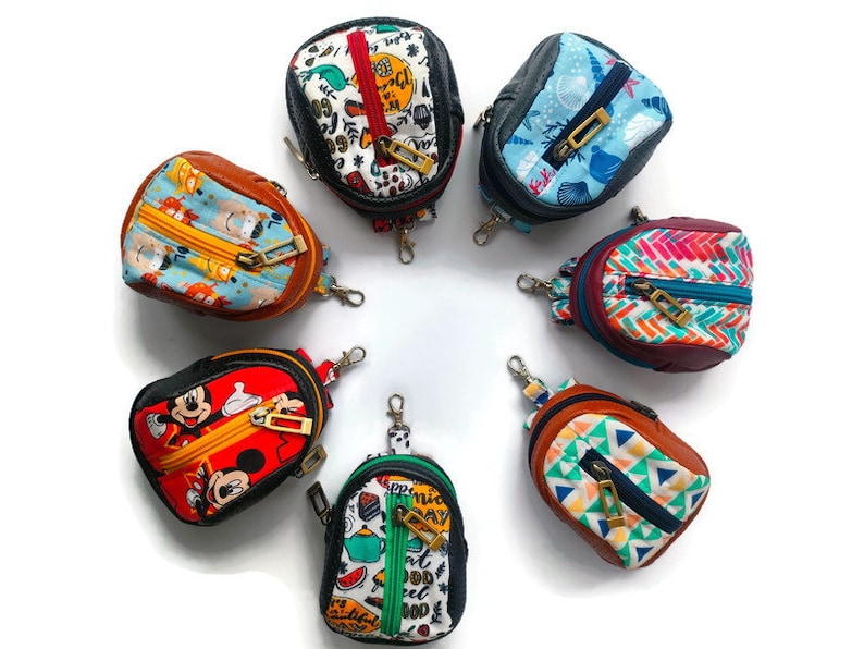 K&ouml;nnte beinhalten: Sieben bunte Mini-Rucks&auml;cke mit Rei&szlig;verschl&uuml;ssen und Schl&uuml;sselanh&auml;ngern. Die Rucks&auml;cke zeigen verschiedene Muster, darunter Comicfiguren, geometrische Designs und abstrakte Kunst.