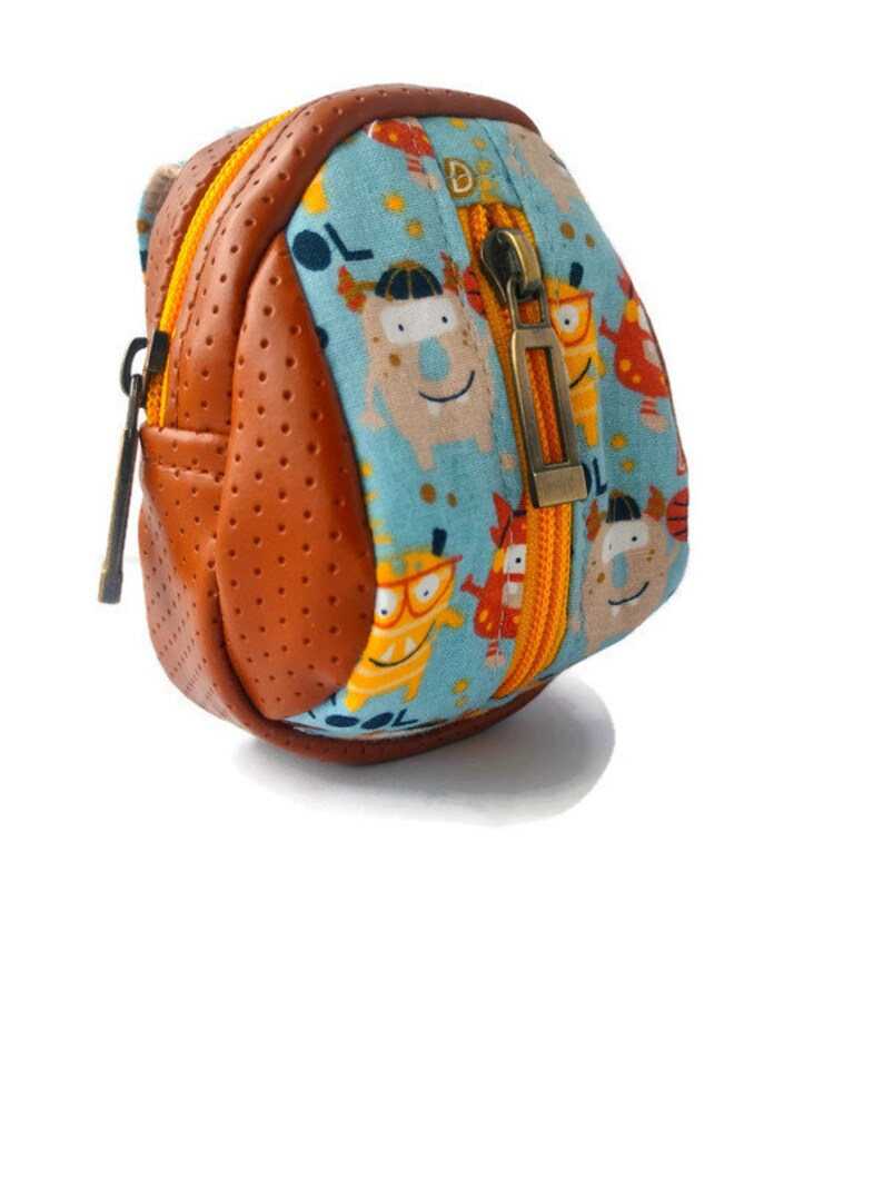 K&ouml;nnte beinhalten: Ein kleiner, brauner und blauer Rucksack mit Rei&szlig;verschluss. Der Rucksack hat einen Cartoon-Monster-Druck mit dem Wort "COOL" darauf.