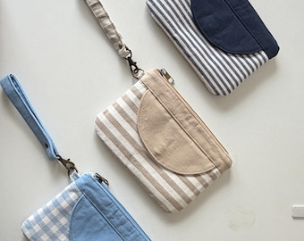 May include: Three rectangular fabric pouches with wrist straps. One pouch is light blue gingham, one is beige and white striped, and one is navy blue and white striped. Each features a solid-colored semi-circular front pocket and a zipper.
