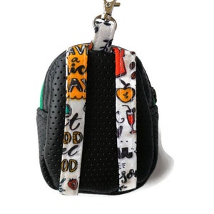 K&ouml;nnte beinhalten: Schwarzer Mini-Rucksack-Schl&uuml;sselanh&auml;nger mit gr&uuml;nem Rei&szlig;verschluss und bunten gemusterten B&auml;ndern. Die B&auml;nder haben den Text "Eat Good Feel Good" und "Have a Nice Day".