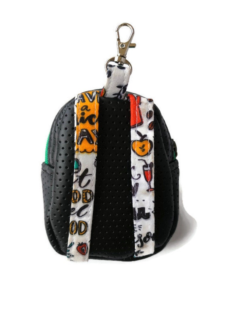 K&ouml;nnte beinhalten: Schwarzer Mini-Rucksack-Schl&uuml;sselanh&auml;nger mit gr&uuml;nem Rei&szlig;verschluss und bunten gemusterten B&auml;ndern. Die B&auml;nder haben den Text "Eat Good Feel Good" und "Have a Nice Day".