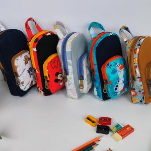 Könnte beinhalten: Ein Set aus sechs kleinen, bunten Rucksäcken mit verschiedenen Comicfiguren darauf. Die Rucksäcke haben Reißverschlüsse und verstellbare Träger.