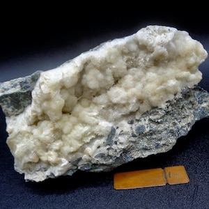 Könnte beinhalten: Nahaufnahme eines weißen und grauen Steins mit einem Cluster aus weißen Kristallen. Der Stein ist etwa 10 cm lang und liegt auf einer schwarzen Oberfläche.