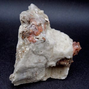 Può includere: Un esemplare minerale naturale e grezzo con una miscela di tonalità bianche, arancioni e marroni. La roccia ha una struttura cristallina con formazioni di cristalli arancioni visibili e una superficie strutturata. L'esemplare è posto su uno sfondo scuro.