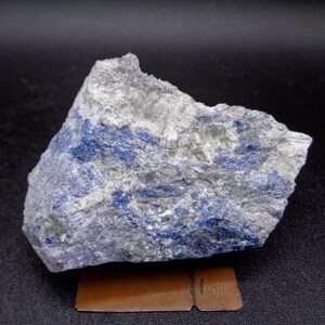 May include: A grey and blue rock specimen with a rough, textured surface. The natural stone has an irregular shape and a mix of colours. A small, brown rectangle with "1 cm" is in the foreground.
