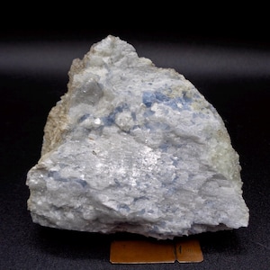 May include: A light grey rock formation with blue inclusions, showing a rough, uneven surface. The mineral specimen has a crystalline texture. The background is dark, highlighting the rock's details.