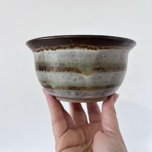 May include: A handmade ceramic bowl with a dark brown rim and a light brown base. The bowl features horizontal bands of green, brown, and white. The bowl is held by a hand against a white background.