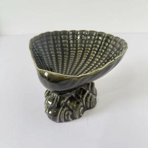 May include: A dark green ceramic seashell-shaped dish. The dish has a textured surface and sits on a decorative base. The seashell bowl is a decorative item.