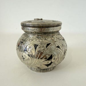 May include: A silver-coloured, lidded container with intricate floral and leaf patterns. The container has a rounded body and a lid with a small loop handle. The metalwork features detailed engravings and a decorative border around the lid and body.