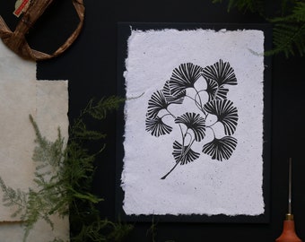 Könnte beinhalten: Schwarz-weiß-Druck eines Ginkgoblatt-Zweigs auf weißem strukturiertem Papier mit schwarzem Rand. Der Druck ist gerahmt und hängt an einer schwarzen Wand.